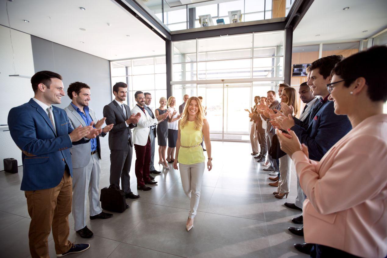 Eine Frau betritt lächelnd ein Bürogebäude, während Kollegen applaudieren. Symbolisiert die positiven Effekte eines Employee Assistance Programs (EAP) auf Wertschätzung und Teamgeist.