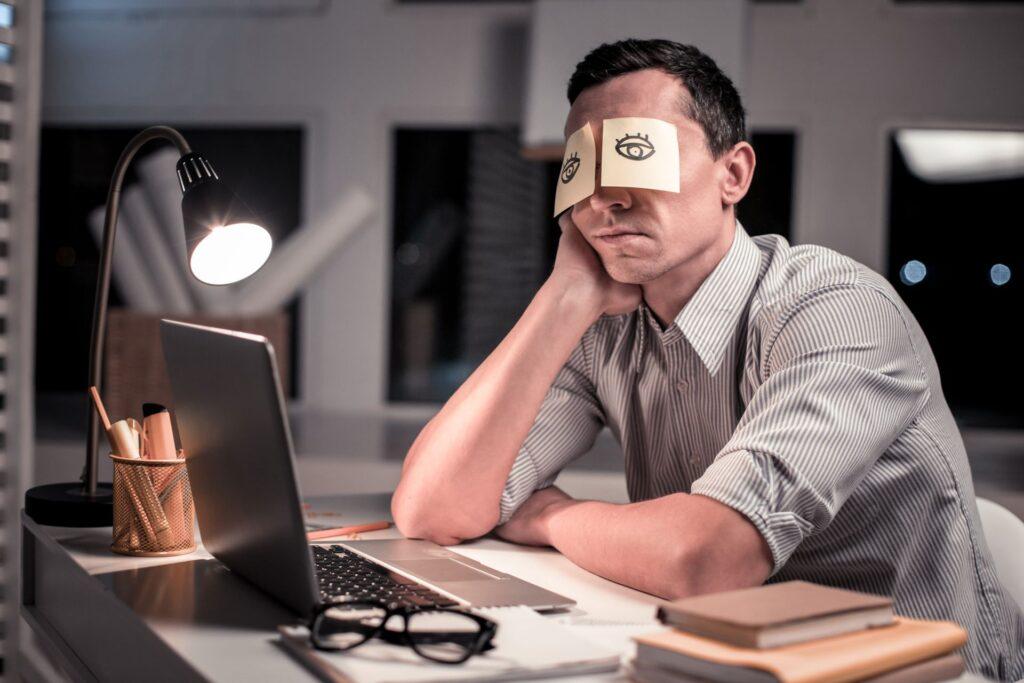 Müder Mann mit Post-its auf den Augen am Schreibtisch